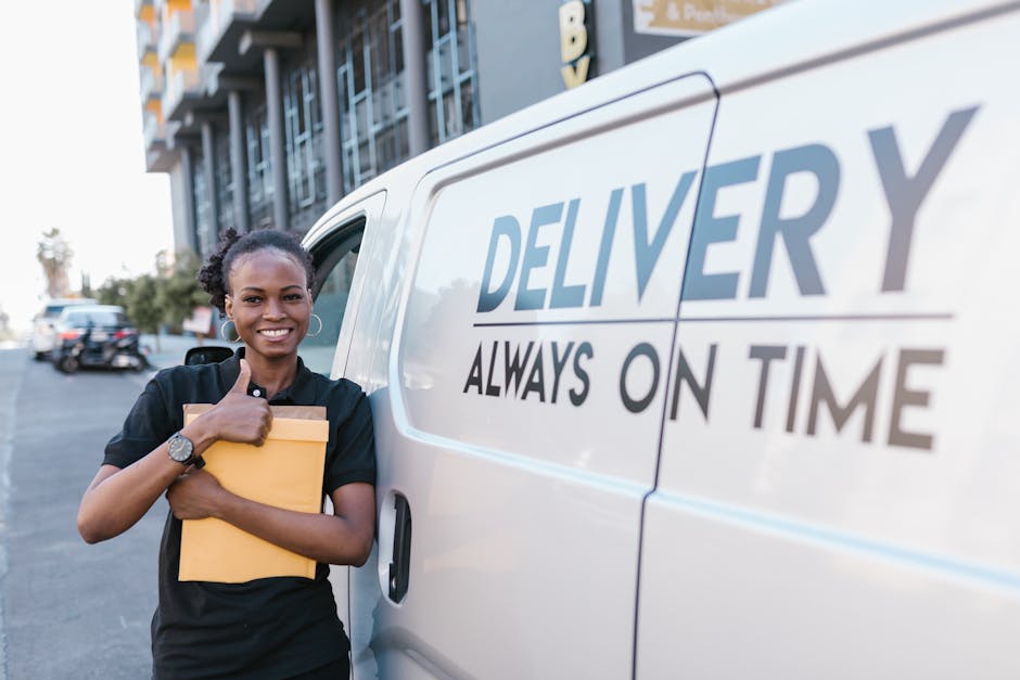 Two movers from Man with Van Thornton Heath are engaged in a furniture transport process, loading a large, dark green upholstered item wrapped in protective fabric into the open rear of a white panel van parked on the street. The van's sliding door is fully open, revealing an interior designed for home relocation tasks, with visible cargo space and securing straps. The movers are standing on the pavement next to the van; one is using both hands to lift and guide the furniture, while the other supports it from the side. They are dressed in dark uniforms with safety wristbands, and the scene is set during daylight under a clear sky. In the background, a street sign indicating Somerset Road is visible, along with other vehicles and urban surroundings. This image illustrates the packing and loading process typical of house removals, showcasing furniture handling and transportation logistics managed by professionals like those from [COMPANY_NAME], supporting efficient moving services in Thornton Heath.