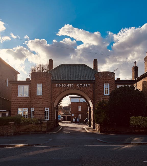 Photograph of the entrance to Knights Court, a red brick residential building with an arched gateway, situated on a street during daytime with partly cloudy skies. The archway leads into a driveway with parked cars visible beyond, and the building features white-framed windows and two chimneys on the roof. Surrounding the entrance are neatly trimmed bushes, and the scene depicts a typical urban environment suitable for home relocations or furniture transport. This setting is associated with property moving services provided by Man with Van Thornton Heath, supporting efficient packing and loading processes during house removals.
