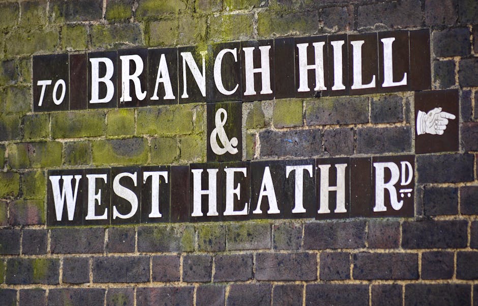 A close-up view of a brick wall with greenish moss growth, displaying a street sign composed of individual black tiles with white uppercase lettering. The sign reads 'TO BRANCH HILL & WEST HEATH RD,' with the words arranged in a staggered layout. To the right of the sign, a small white ceramic tile features a detailed illustration of a pointing hand. The overall scene depicts an outdoor setting, likely on a residential or commercial property, with the sign indicating directions for a home relocation or moving process. The image reflects elements related to house removals and transport logistics, relevant to services offered by Man with Van Thornton Heath, especially concerning local street signage in Croydon.