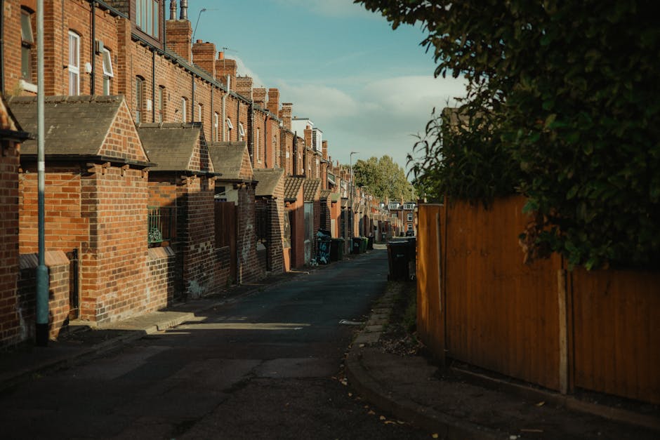 The image shows a narrow residential street in Thornton Heath, featuring rows of brick terraced houses with small front gardens and pitched roofs. The street is lined with parked wheelie bins and trash containers near the pavement, indicating waste collection. On the right side, a tall wooden fence runs alongside a hedge, partially obscuring the view of the houses. The asphalt road appears to be clear, with some shadows cast by nearby structures and trees. The scene is illuminated by natural daylight, with a partly cloudy sky visible overhead. This setting reflects a typical London suburb environment suitable for house removals or moving logistics, as provided by local services such as Man with Van Thornton Heath. Such areas are common for home relocation, furniture transport, and packing and moving activities, with ample space for loading and unloading furniture and moving boxes.
