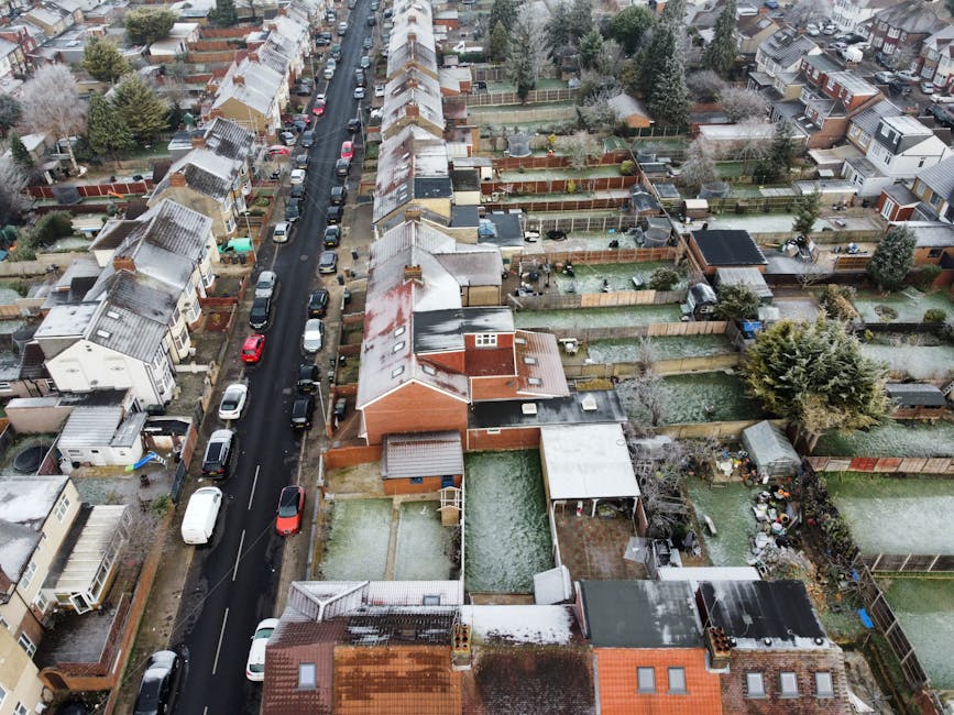 An aerial view of a residential street in Croydon during winter, showing a row of terraced houses with snow-covered roofs and gardens. Several cars are parked along the narrow, asphalt road, which runs down the center of the image. Backyard fences, some wooden and others brick, enclose various garden spaces, with a few containing garden furniture, sheds, or small patios. The scene includes trees, some evergreen, with snow on their branches, and the overall area appears quiet and well-maintained. The image captures the typical urban environment involved in house removals or home relocation services. Occasionally, the logistics of loading or unloading might be inferred from the scene's arrangement, relevant to companies like Man with Van Thornton Heath and their house clearance or furniture transport services. The overall setting highlights the urban residential environment where efficient packing and moving are essential for complying with local house clearance laws in Croydon, with careful attention to outdoor space preparation for home relocation activities.