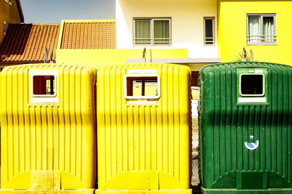 An outdoor area filled with large stacks of flattened cardboard boxes displaying fresh fruit branding, positioned behind metal platform carts loaded with smaller boxes and packaged items, alongside several plastic storage bins in green and red. The scene shows the process of handling recyclable materials and packing supplies in preparation for a house removal or home relocation. In the background, there is a residential neighborhood with trees and rooftops visible under daylight. The setting appears to be adjacent to a property, on a paved surface, with a focus on the organization and transportation of packing materials. This image illustrates the logistics involved in packing and moving processes, as managed by companies like Man with Van Thornton Heath, who specialise in removals and waste disposal services in Thornton Heath.