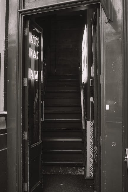 A black and white photograph showing a narrow staircase inside a building, viewed from the bottom looking up through an open doorway with a wooden frame. The staircase has dark wooden steps and a simple, utilitarian design, with no visible handrails. To the left side of the doorway, there are partially visible stickers or signs with text, partially obscured by the door frame. The surrounding interior walls appear dark and plain. This scene likely depicts a typical house or apartment interior used in a home relocation or moving process, consistent with services offered by Man with Van Thornton Heath, specializing in removals and furniture transport. The lighting appears to come from natural light filtering in from the top of the staircase, illuminating the steps and creating a contrast with the darker areas around the door frame.