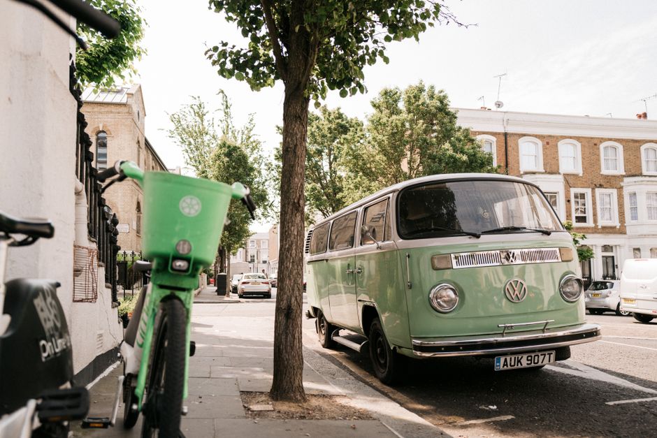 A vintage light green Volkswagen Type 2 van parked on the street near a tree on London Road, Thornton Heath, during daylight hours. The van's side sliding door is closed, and it is positioned adjacent to the pavement, with a black wheel visible at the front. In the foreground, part of a green bicycle with a front basket is seen leaning against a wall, while a bicycle lock is attached nearby. In the background, several cars are parked along the street, with residential buildings featuring bay windows and brick facades visible behind. The scene is well-lit with natural sunlight, and the sidewalk appears to be used for facilitating home relocations and moving logistics, with the van likely loaded or preparing for furniture transport, as part of the local removals services provided by Man with Van Thornton Heath.