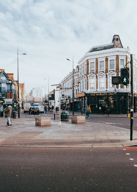 A view of Thornton Heath High Street during daytime, featuring a busy street scene with pedestrians walking on the pavement, some carrying shopping bags, and vehicles including a white van and several cars moving along the road. On the right side, there is a four-story corner building with a white facade, large windows, and a sign for 'The World's End' pub at ground level. To the left, there are shops and storefronts, with outdoor seating areas and people standing near the storefronts. The street is equipped with traffic lights, including a green pedestrian light, and traffic signs. The sky is partly cloudy, and the environment suggests a typical urban area suitable for house removals and relocation services, such as those provided by Man with Van Thornton Heath.