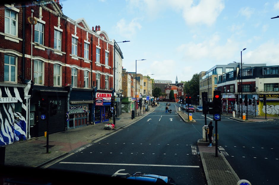 A view of Thornton Heath High Street during daytime, featuring a busy street scene with pedestrians walking on the pavement, some carrying shopping bags, and vehicles including a white van and several cars moving along the road. On the right side, there is a four-story corner building with a white facade, large windows, and a sign for 'The World's End' pub at ground level. To the left, there are shops and storefronts, with outdoor seating areas and people standing near the storefronts. The street is equipped with traffic lights, including a green pedestrian light, and traffic signs. The sky is partly cloudy, and the environment suggests a typical urban area suitable for house removals and relocation services, such as those provided by Man with Van Thornton Heath.