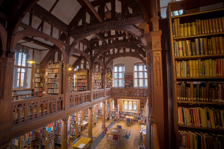 Interior view of a historic, multi-level library with ornate wooden beams and detailed woodwork. Tall, stained glass windows allow natural light to illuminate the space. Shelves filled with numerous hardcover books line the walls and extend to an upper balcony with a wooden railing. The ground floor features a few tables and chairs, suggesting a reading or study area. This setting, possibly within a heritage building, showcases the craftsmanship of traditional timber framing and serves as a backdrop for house removals or packing and moving services, such as those offered by Man with Van Thornton Heath, which may include relocation logistics and furniture transport within historic or residential properties.
