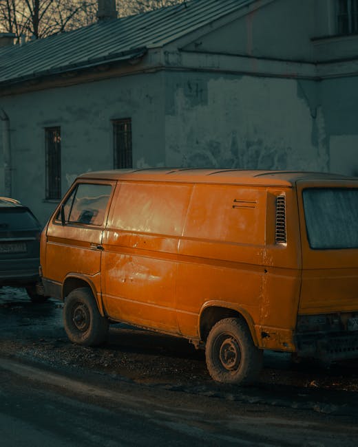 A weathered orange van parked on a street near a white building with small, rectangular windows. The van's exterior shows signs of rust and dirt, and it is positioned close to the kerb, with its rear facing a narrow road. To the left of the orange van, a dark grey car is partially visible, parked alongside the van. The scene is illuminated by natural daylight, highlighting the aged condition of the vehicle. This setting captures a typical urban street scene, suitable for house removals or furniture transport services. Occasionally, Man with Van Thornton Heath’s home relocation team might use such vehicles to assist with loading household furniture and packed boxes into the van for efficient moving logistics, often involving careful handling, packing materials like cardboard boxes and plastic wrap, and loading processes designed to ensure safe transport of belongings during a house move near Thornton Heath Pond.